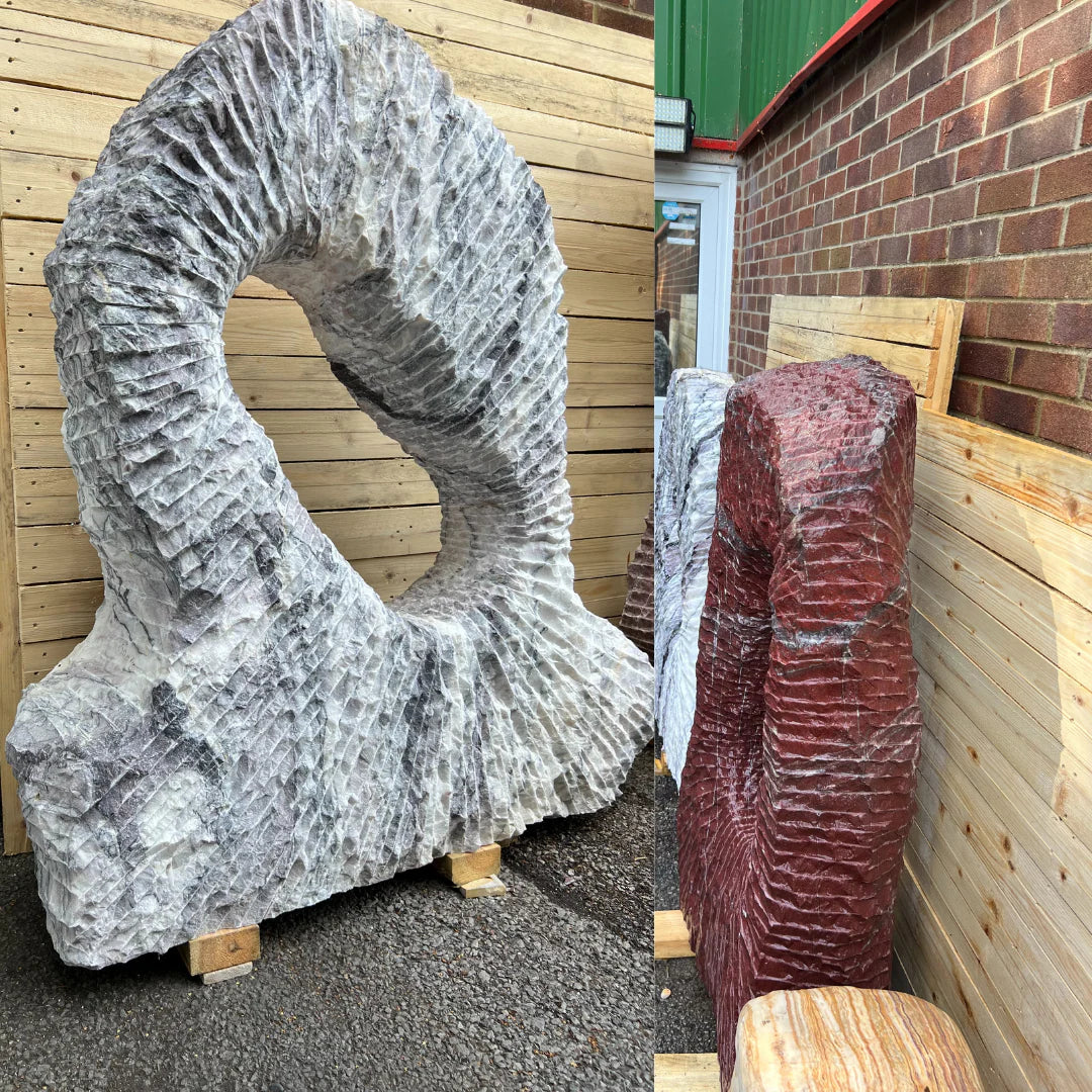 Circular Leylak natural stone monolith with a sculpted hollow center, featuring textured grey and purple tones, displayed outdoors beside red decorative stone pieces at Stones4Gardens.